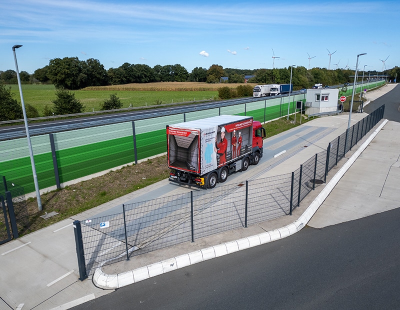LKW von Pfreundt auf Straße, umgeben von Feldern und Windrädern, zeigt Werbung für mobile Wiegesysteme auf Plane.