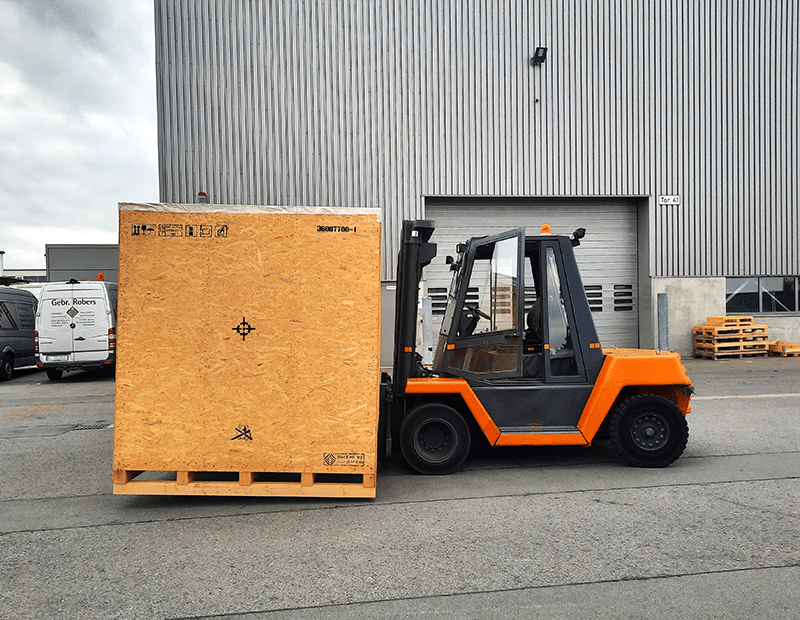 Gabelstapler hebt große Holzkiste vor Lagerhalle, geeignet für Pfreundts mobile Wiegesysteme wie Radladerwaagen.