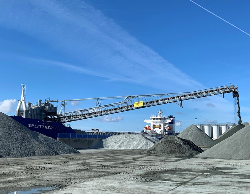 Industriegelände mit Förderband und Frachtschiff, Beladung von Schüttgut im Hafen; blauer Himmel im Hintergrund.