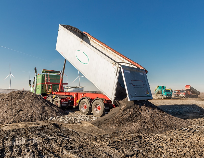 Lkw entlädt Erde auf Baustelle unter blauem Himmel, Windkraftanlagen im Hintergrund.