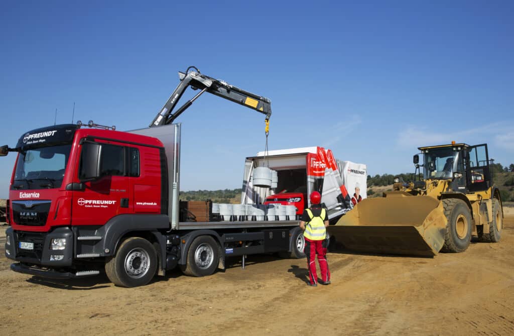 Roter Pfreundt-LKW mit mobilen Wiegesystemen, Bagger und Arbeiter in Schutzkleidung auf Baustelle bei sonnigem Wetter.