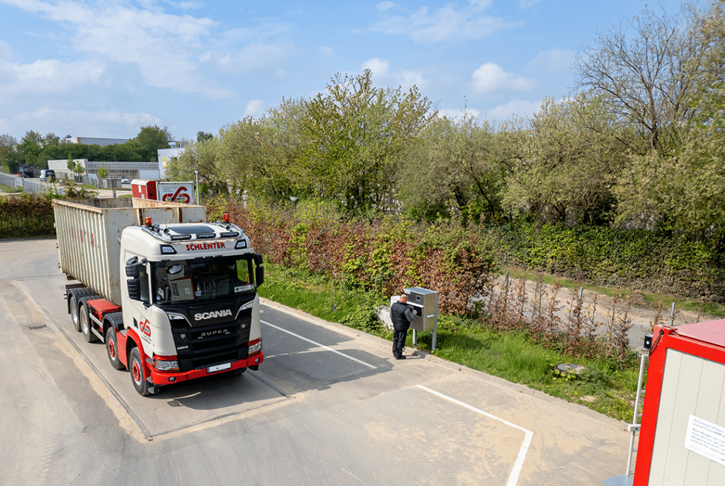 LKW auf Pfreundt-Waage mit Bediener an Kontrollstation, umgeben von grüner Landschaft und blauem Himmel.