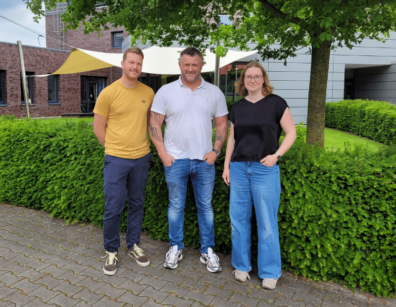 Drei Personen vor einem Firmengebäude von Pfreundt, Anbieter von mobilen Wiegesystemen, lächeln in die Kamera.