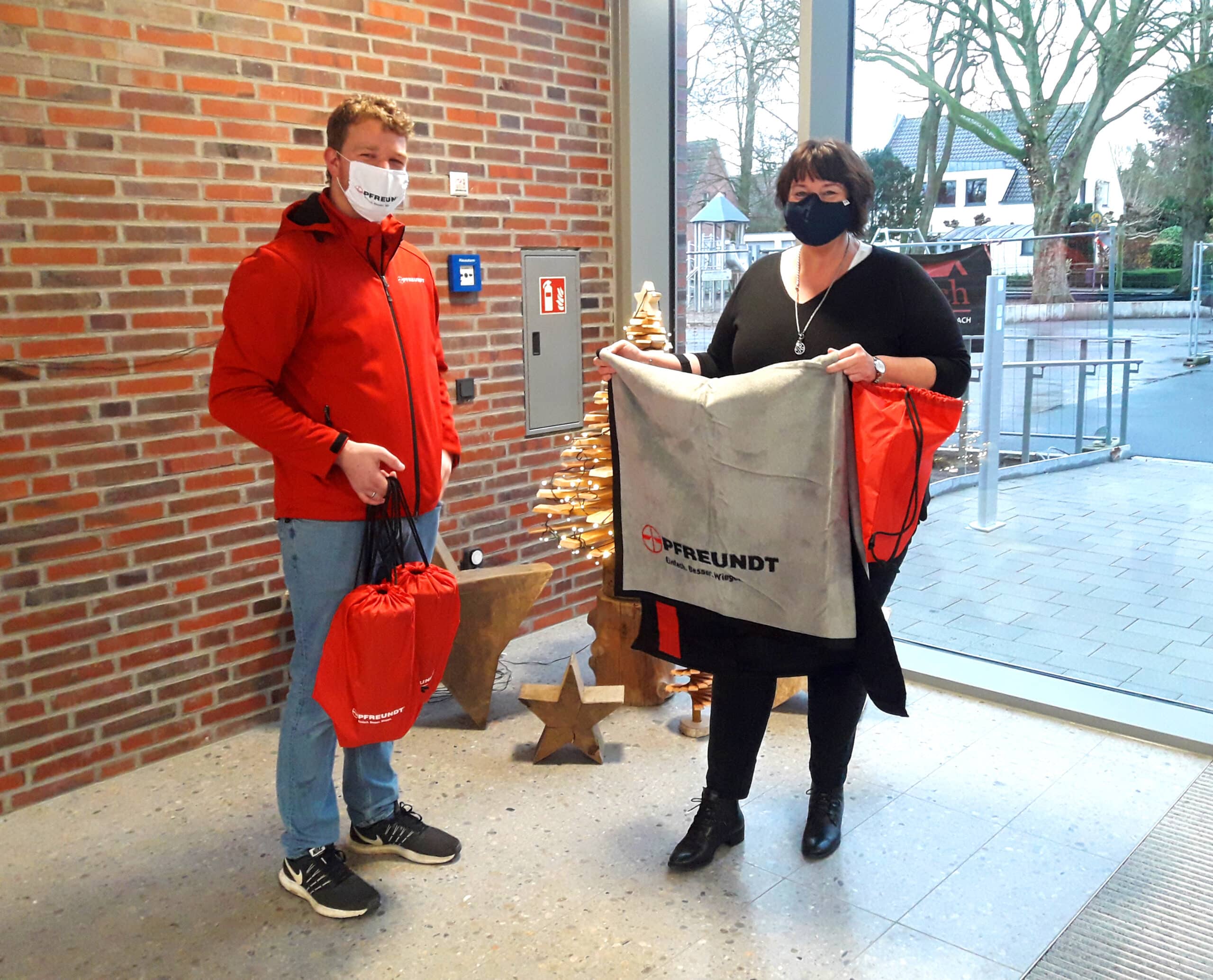 Zwei Personen mit Pfreundt-Werbeartikeln vor einem Weihnachtsbaum in einem modernen Bürogebäude.