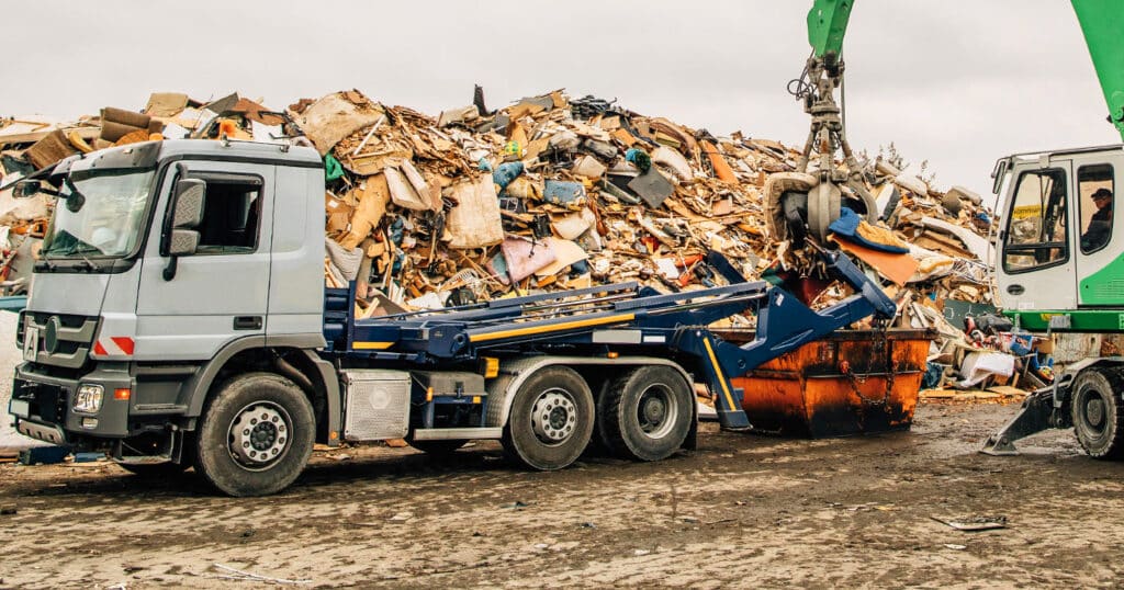 LKW lädt Schrott mit Bagger auf, im Hintergrund großer Schrotthaufen, Nutzung von Pfreundt Wiegesystemen.