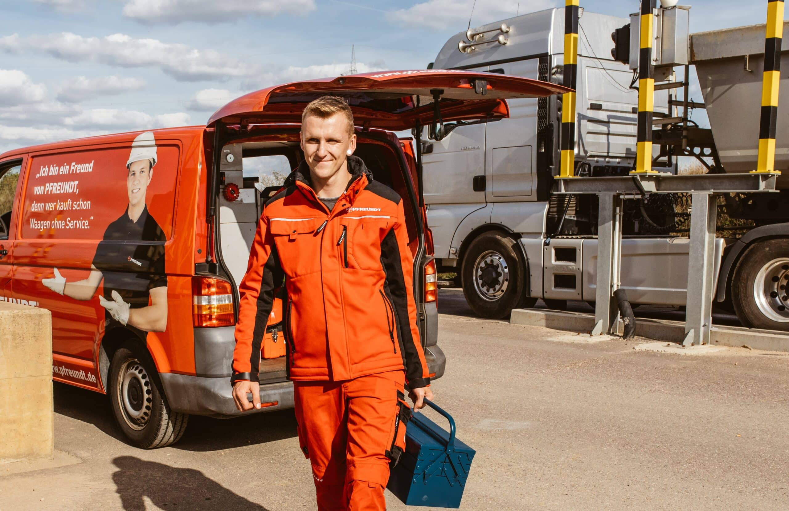 Pfreundt-Mitarbeiter in rotem Anzug mit Werkzeugkoffer vor Firmenfahrzeug und LKW, spezialisiert auf mobile Wiegesysteme.