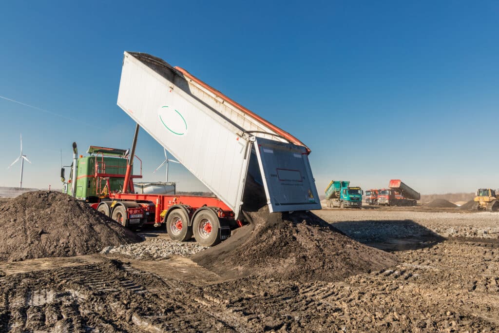 Lkw beim Entladen von Erde auf Baustelle, umgeben von weiteren Fahrzeugen und Windkraftanlagen unter klarem Himmel.