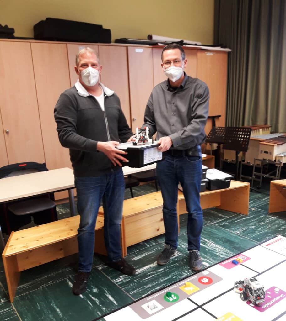 Zwei Personen mit Masken präsentieren ein technisches Gerät in einem Besprechungsraum.