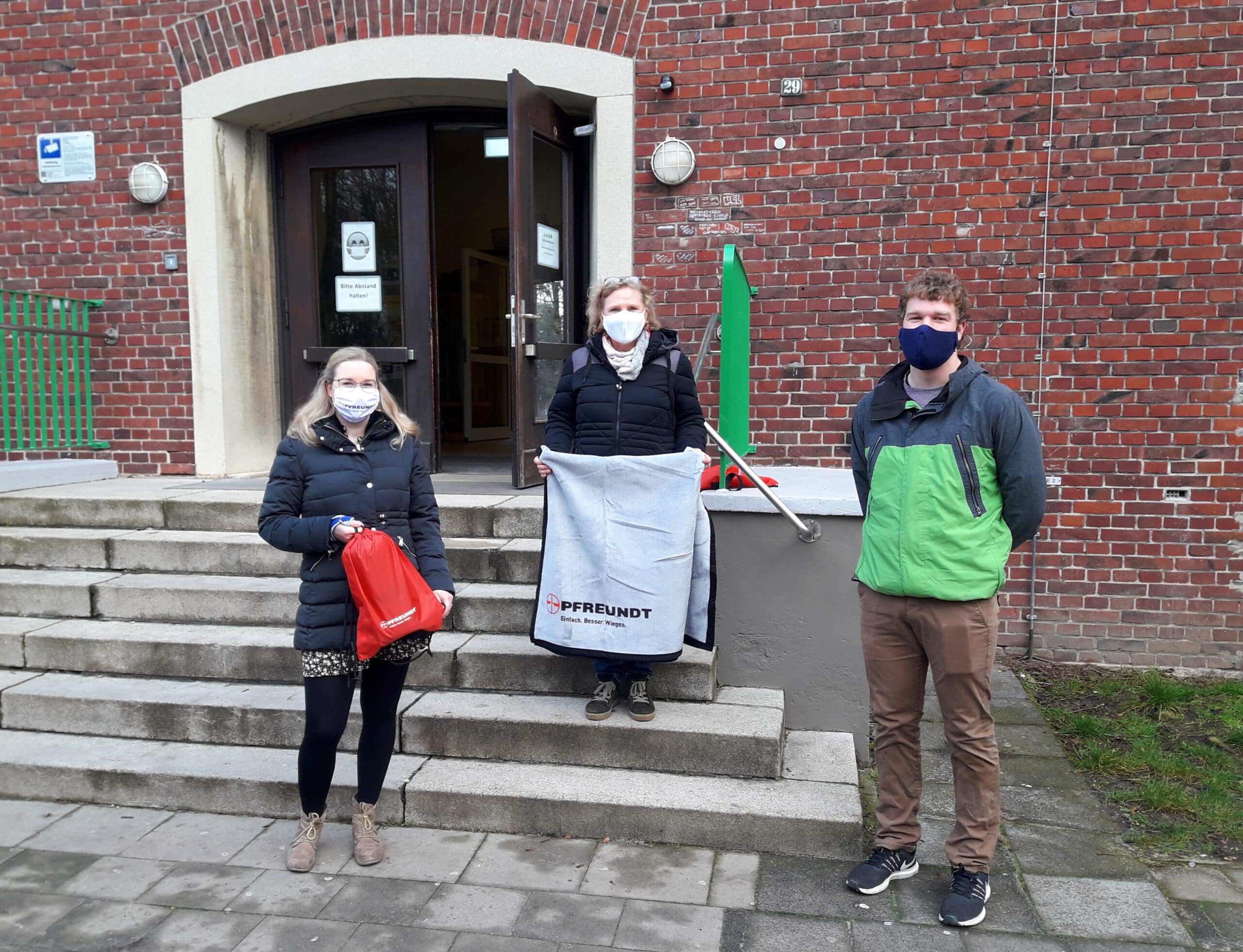 Drei Personen halten Pfreundt-Werbematerial vor einem Backsteingebäude und tragen Masken, stehend auf Treppenstufen.