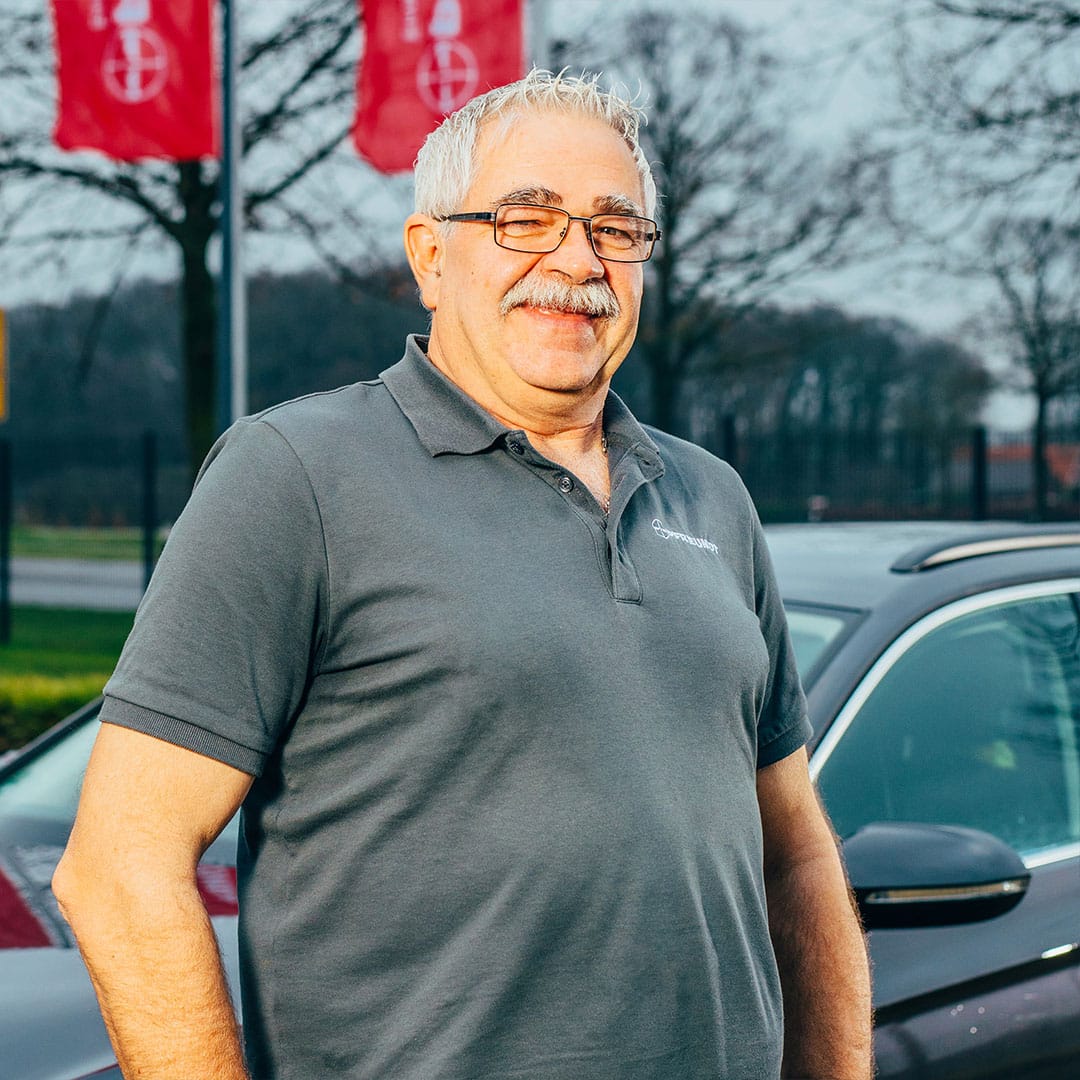 Älterer Herr in grauem Pfreundt-Polo steht vor Fahrzeug, mit Pfreundt-Flaggen im Hintergrund.