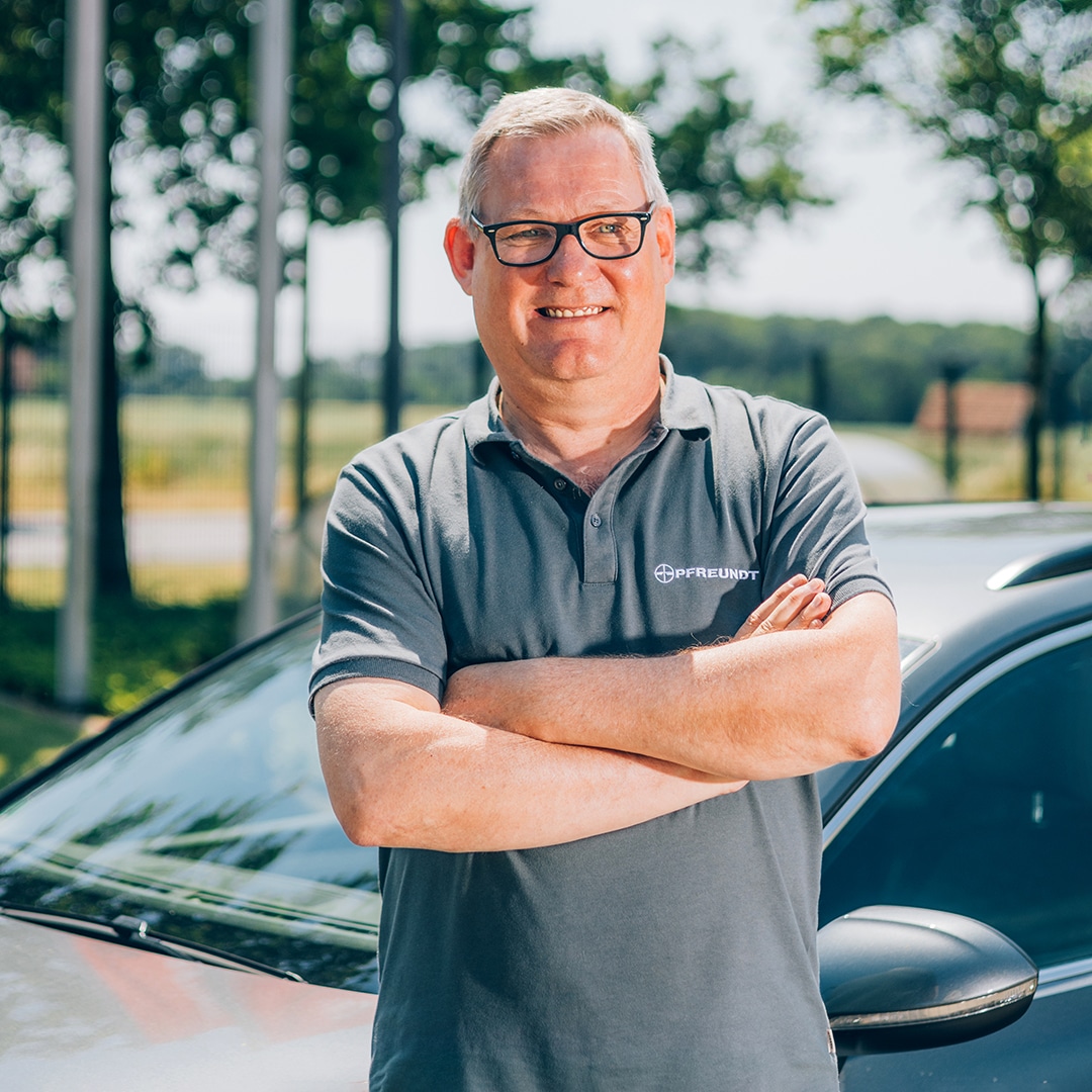 Mann in Pfreundt-Polohemd lächelt vor Auto im Freien. Pfreundt bietet mobile Wiegesysteme für verschiedene Anwendungen.