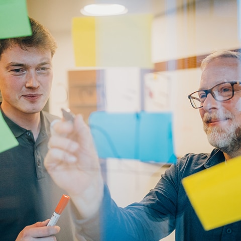 Zwei Männer arbeiten mit bunten Haftnotizen und Stift an einem Whiteboard bei Pfreundt, Anbieter mobiler Wiegesysteme.