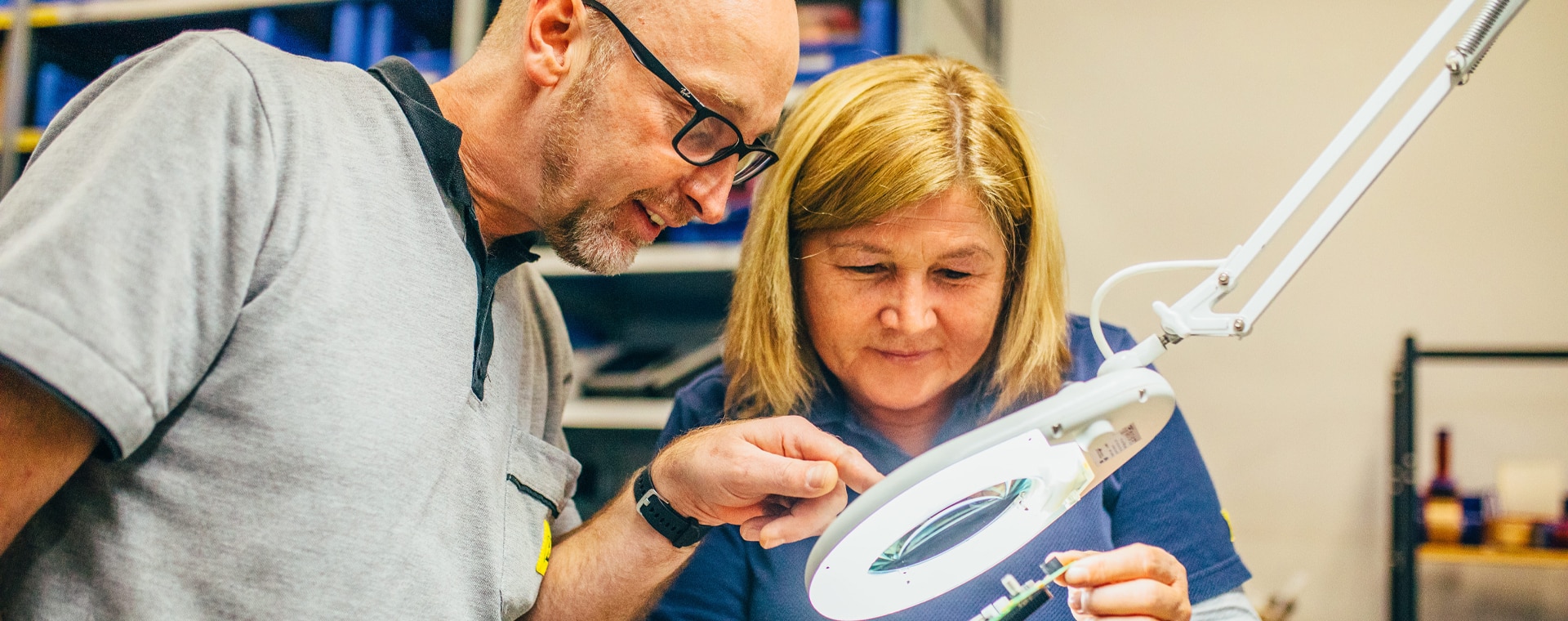 Zwei Personen arbeiten konzentriert an einem Elektronikbauteil unter einer Lupenlampe in einem technischen Labor.